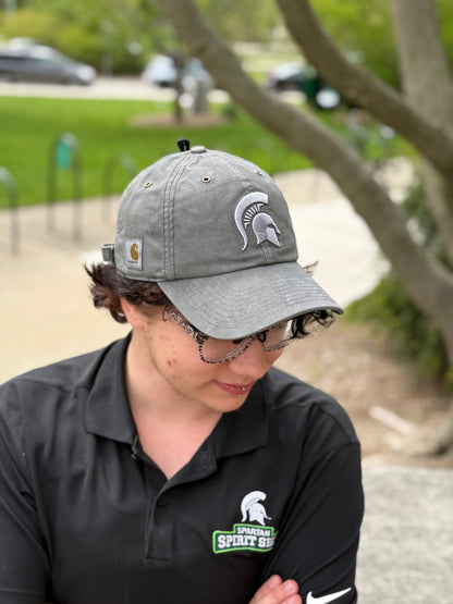 Person standing outdoors showing a gray hat with a light gray Spartan Helmet logo embroidered on the front and Carhartt logo patch on the right side. 