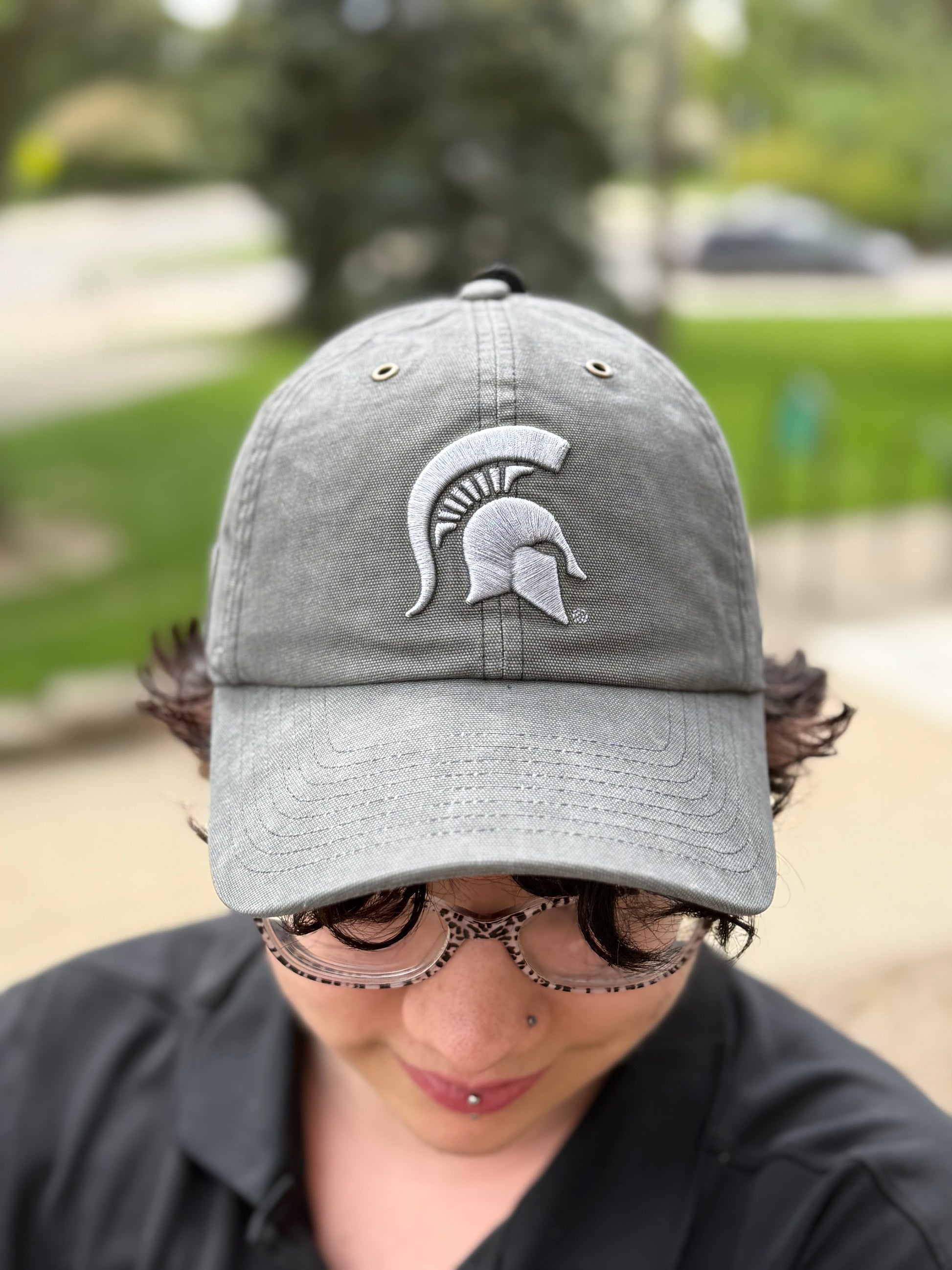 Person standing outdoors showing a gray hat with a light gray Spartan Helmet logo embroidered on the front. 