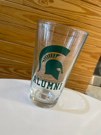 Clear pint glass with a green logo and green Spartan helmet and green 'Alumni' text underneath.