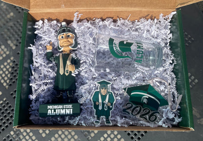 Graduation-themed set with a glass, bobblehead, Grad Sparty sticker, 2026 grad cap MSU ornament with a 'Congrats Graduate' green box.