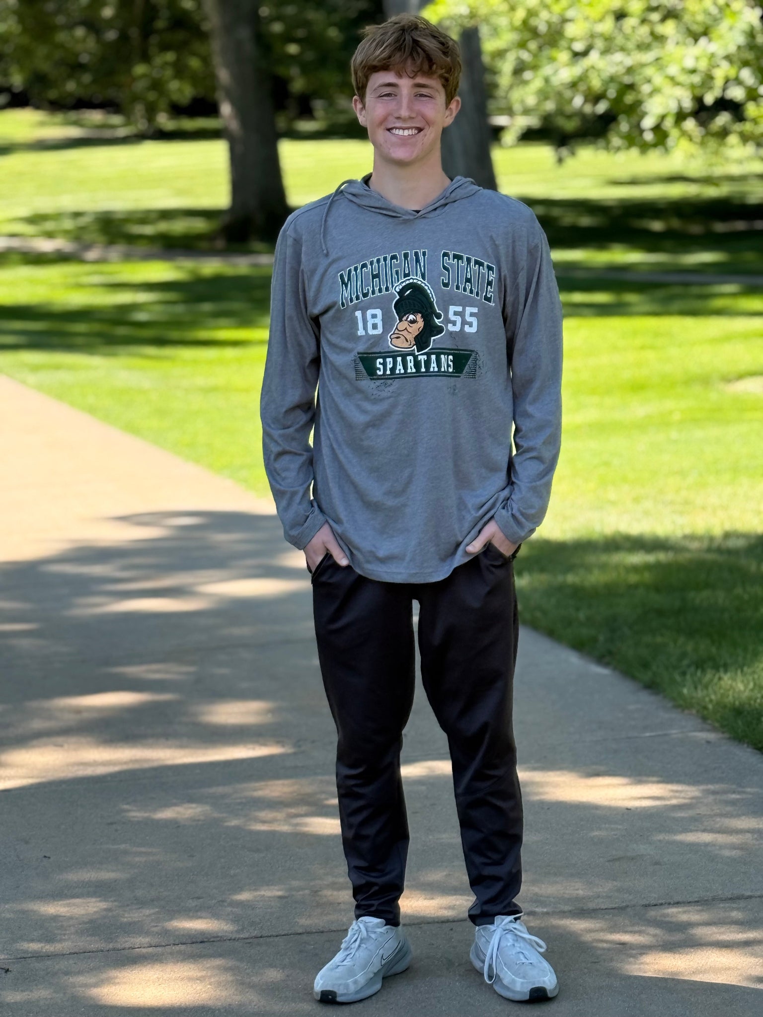 Person wearing gray hooded long sleeve shirt. Center on shirt is logo of gruff sparty with 18 on the left and 55 on the right.  Above Gruff is Michigan State in green and outlined in white. Under gruff sparty is a green banner with Spartans in white. 