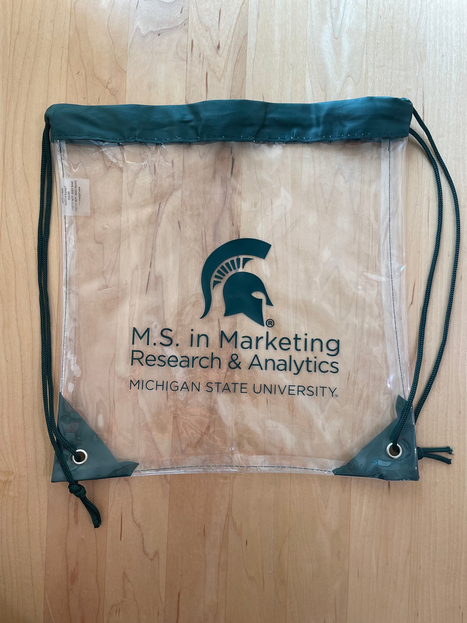 Clear drawstring backpack with the M.S. in Marketing Research & Analytics Michigan State University logo printed in green. Green trim and string handles. 
