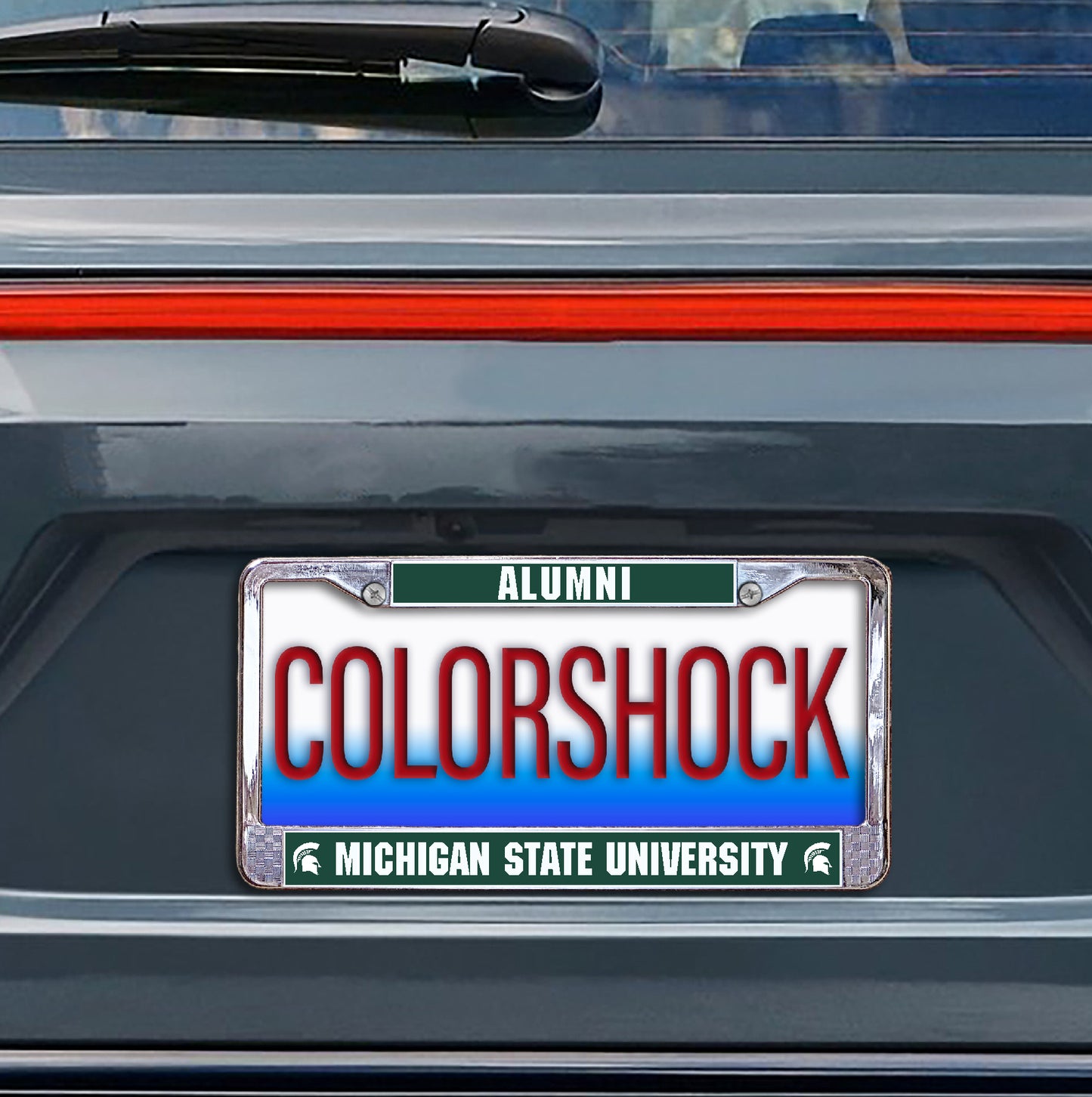 Green and silver license plate with "Alumni" written across the top and "Michigan State University" written across the bottom. Shown on the black of a car. 