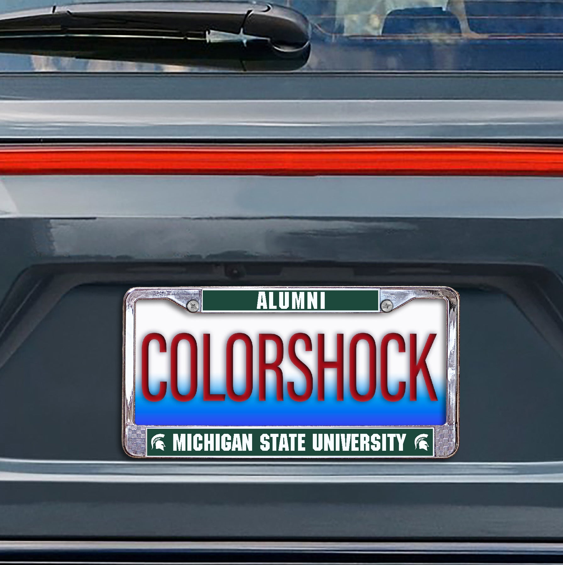 Green and silver license plate with "Alumni" written across the top and "Michigan State University" written across the bottom. Shown on the black of a car. 