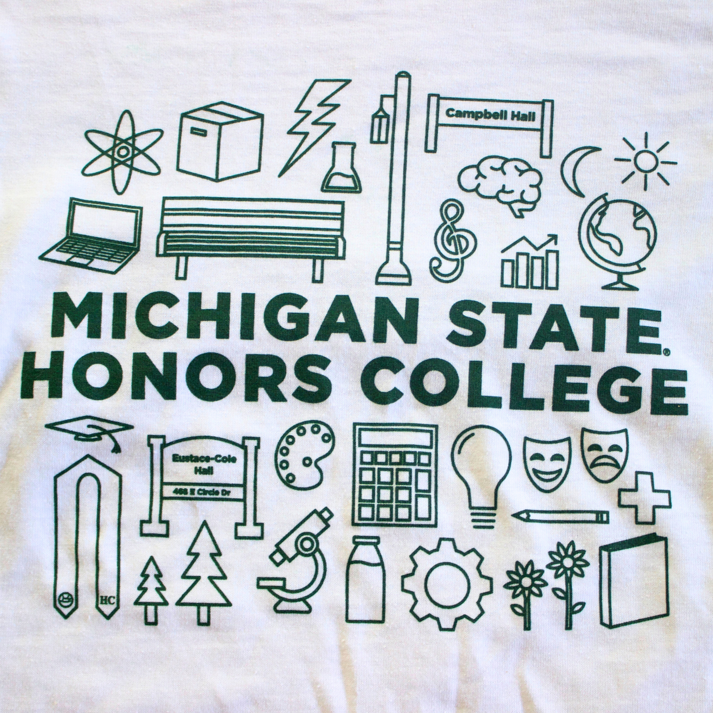A close up of a white T-shirt featuring various forest green icons representing Honors College student interests, studies, and landmarks. The icons include symbols such as a graduation cap, a bench, a globe, a light bulb, theatrical masks, and a tree. The forest green text in the middle of the icons reads: "Michigan State Honors College."