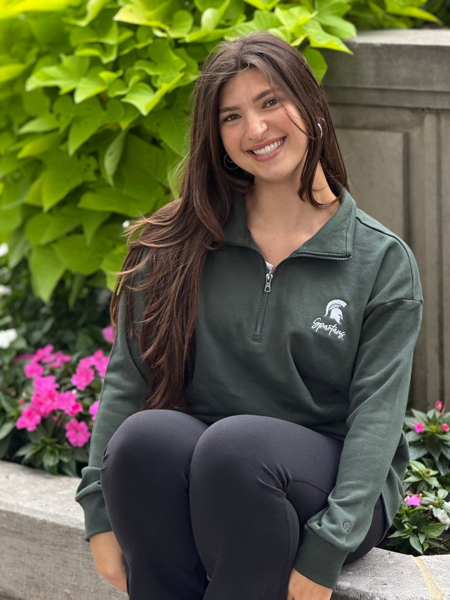 Woman wearing a green sweatshirt with a logo, sitting outdoors with greenery and flowers in the background.