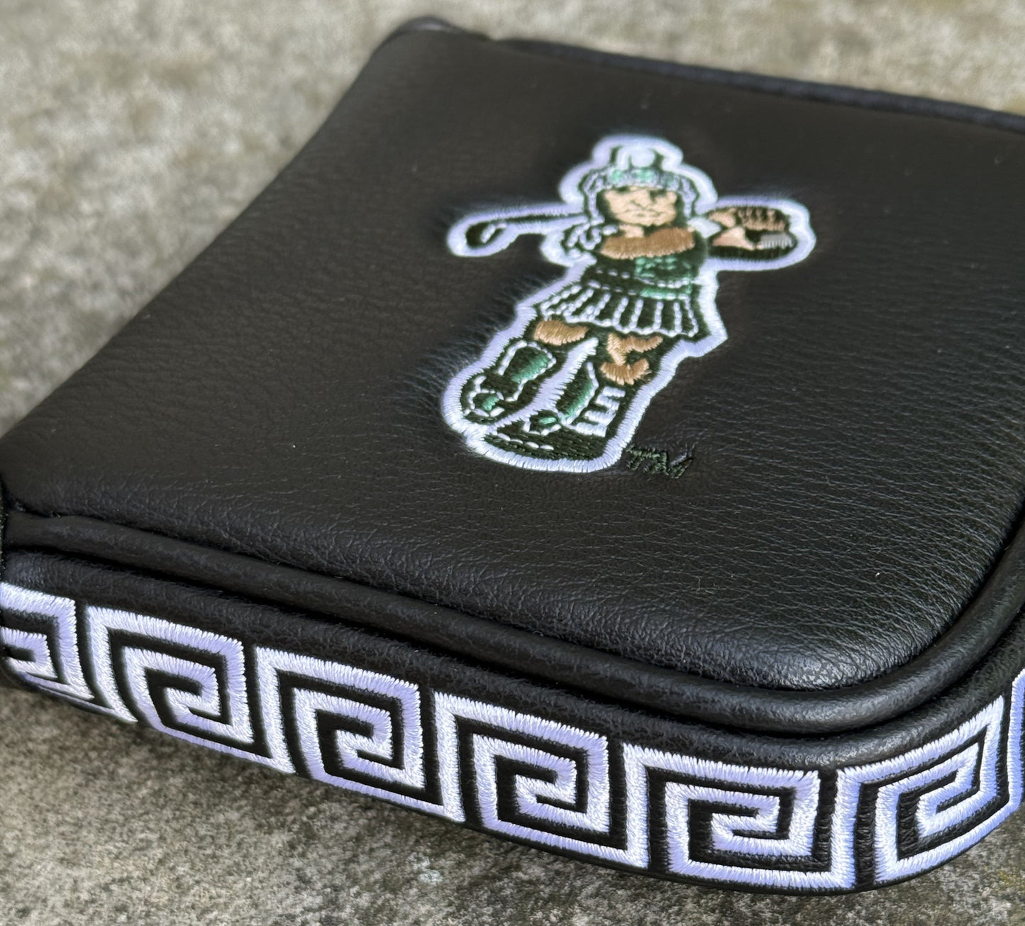 Close up of a black mallet putter headcover with an embroidered Golf Sparty logo on the front and a green and white Greek design on the side. 