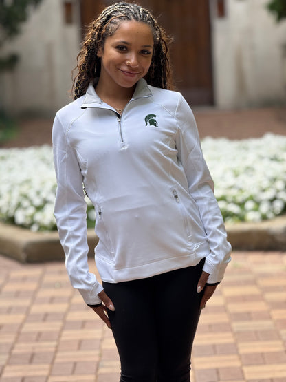 Woman, standing outdoors, wearing a white golf quarter-zip pullover with a green Spartan Helmet logo on the left side of the chest. The quarter-zip has a pocket in the middle with zippers on each side. 