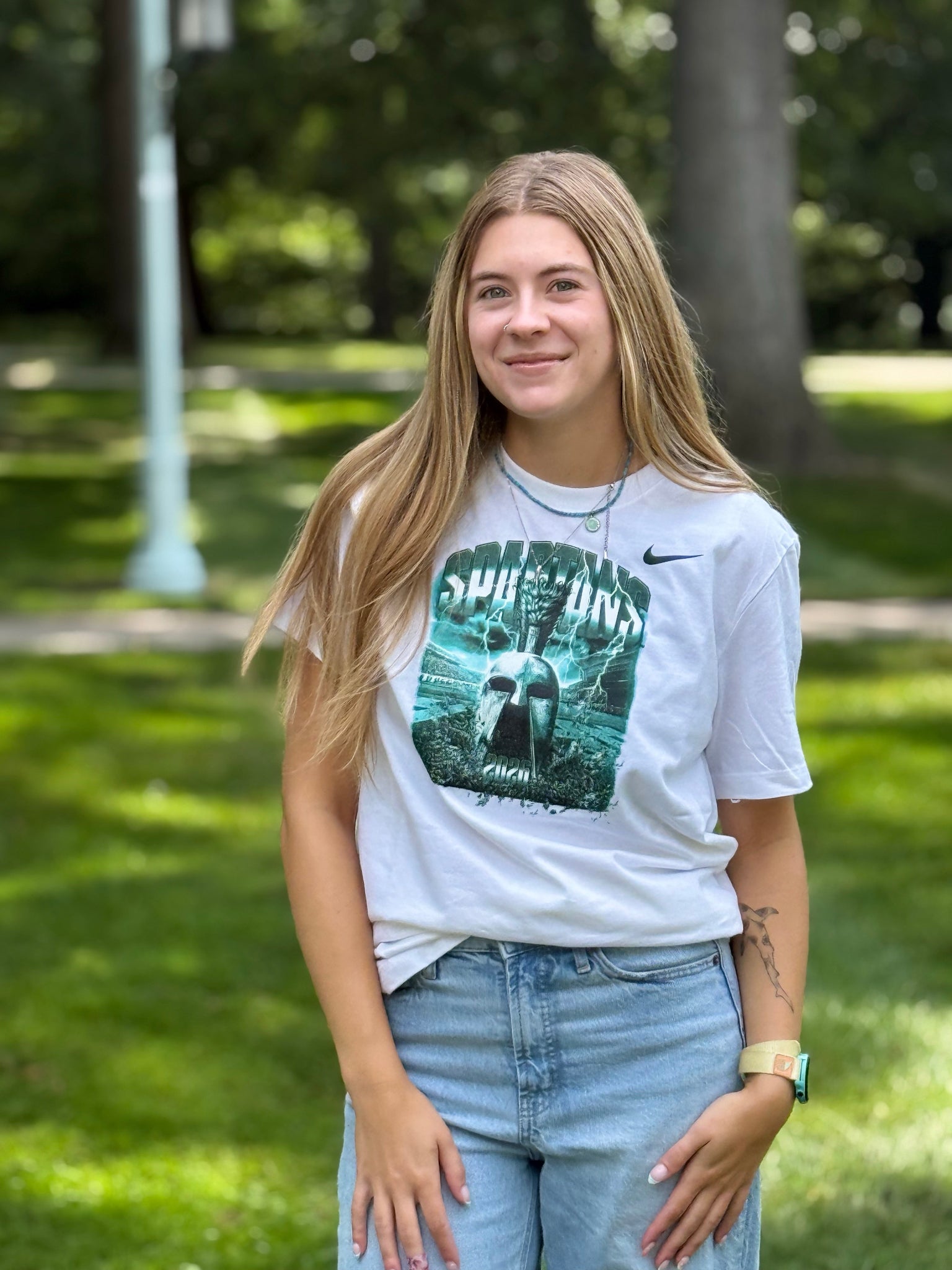 Person standing outdoors on a grassy campus area wearing a white Nike Michigan State Spartans graphic T-shirt featuring a large Spartan helmet design in green tones and light-wash jeans. Trees and a walkway are softly blurred in the background. The shirt displays “SPARTANS” across the chest above the graphic artwork.