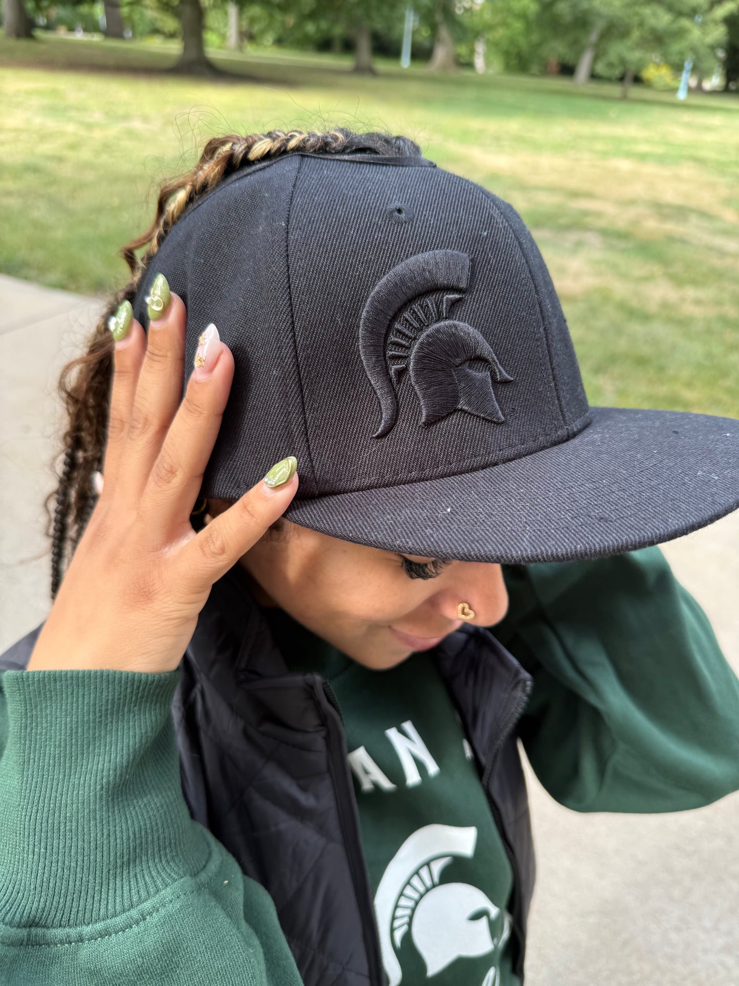 Woman standing outdoors, wearing a black flat-brim cap with an embossed Spartan helmet logo on the front panel. The top of the hat has an opening at the top to protect your hair.