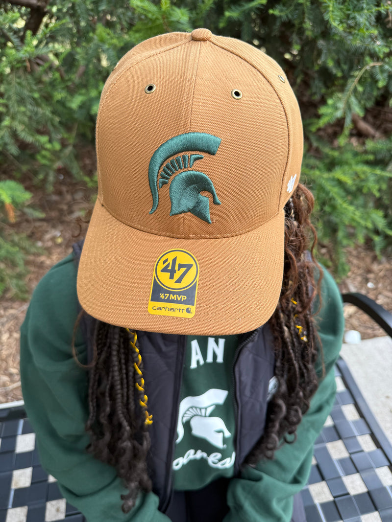 Person wearing a tan cap with a logo and text, sitting outdoors.