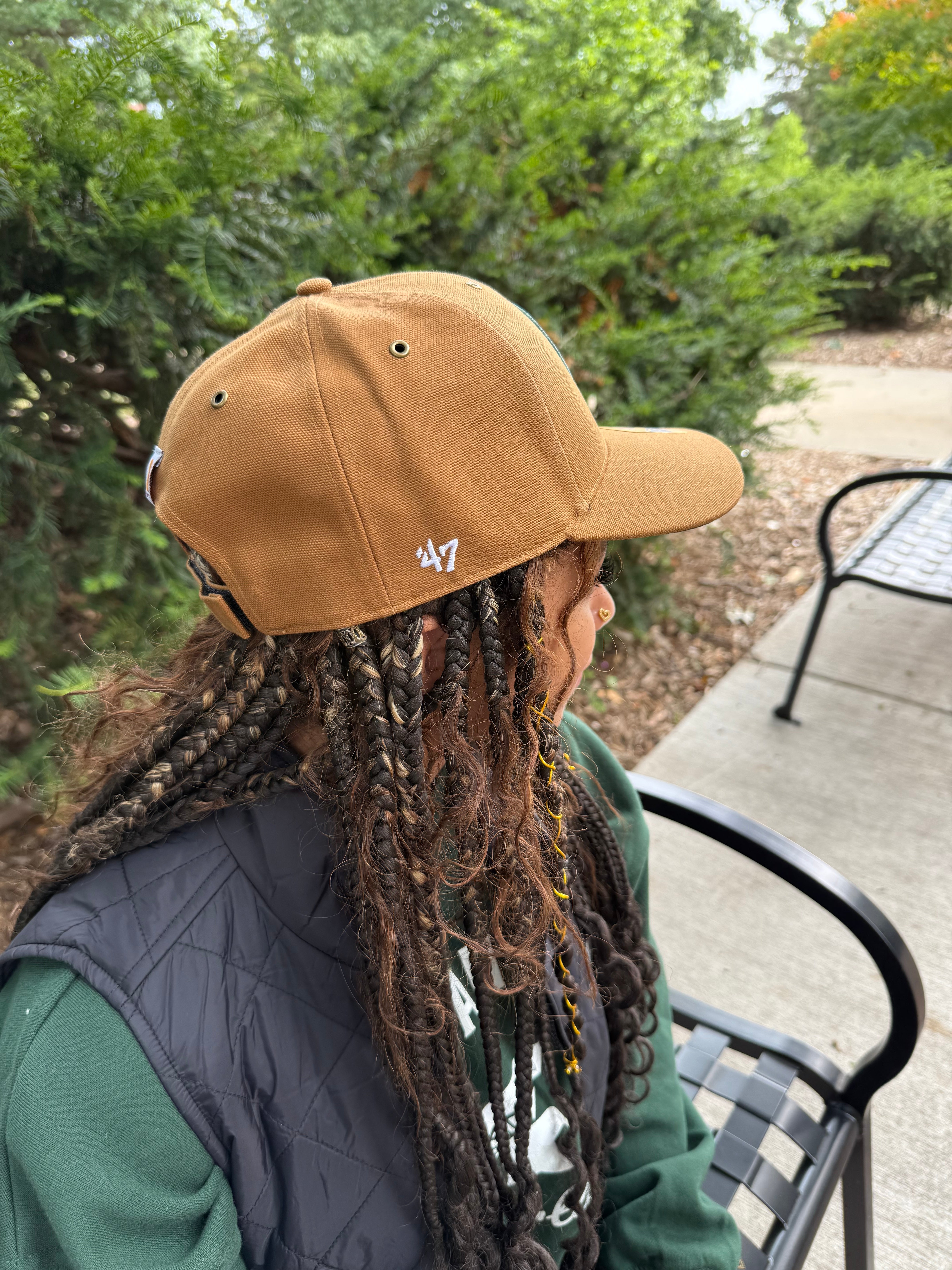 Person wearing a tan cap with a logo, sitting on a bench outdoors.