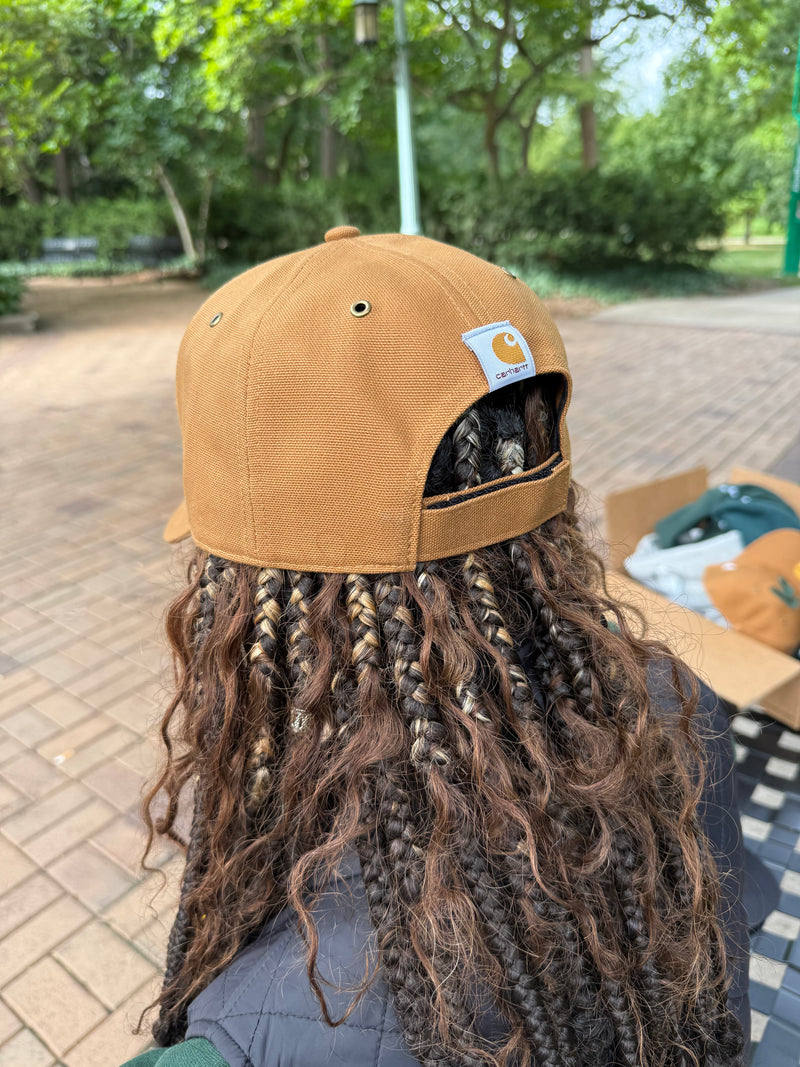 Brown cap with a visible brand logo worn by a person with long, braided hair outdoors.
