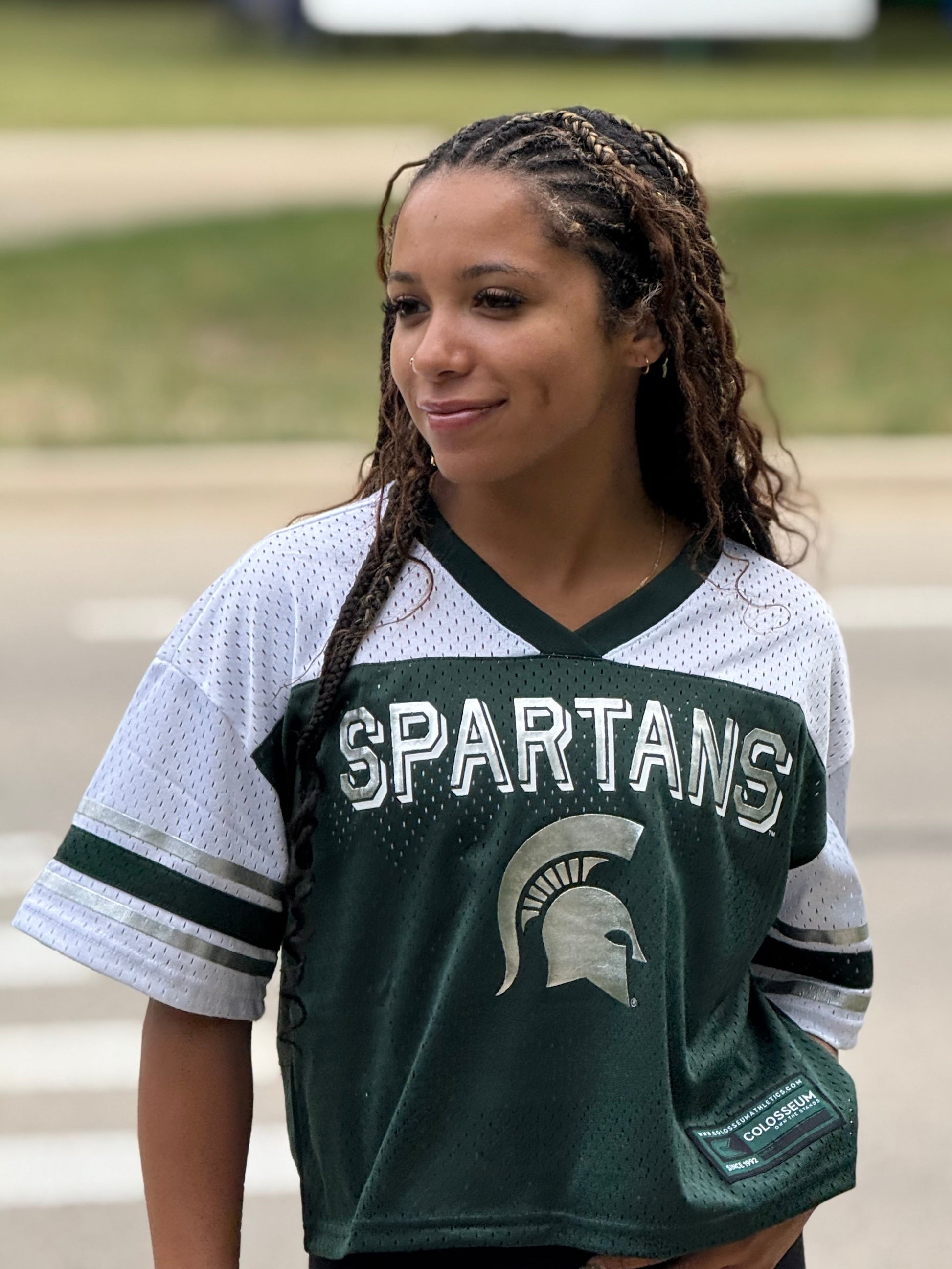 Person, standing outdoors, wearing a Spartans jersey with white short sleeves, a silver Spartans wordmark across the chest, and a silver Spartan helmet logo printed below. 