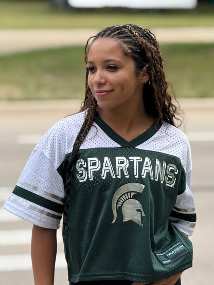 Person, standing outdoors, wearing a Spartans jersey with white short sleeves, a silver Spartans wordmark across the chest, and a silver Spartan helmet logo printed below. 