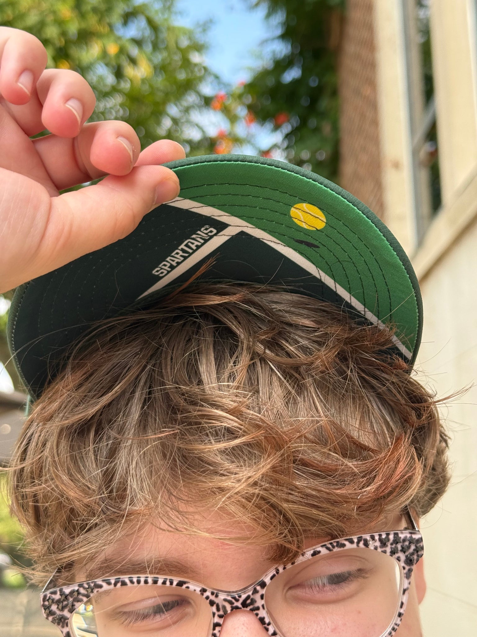 Person holding a green cap with a tennis court design underneath the bill.