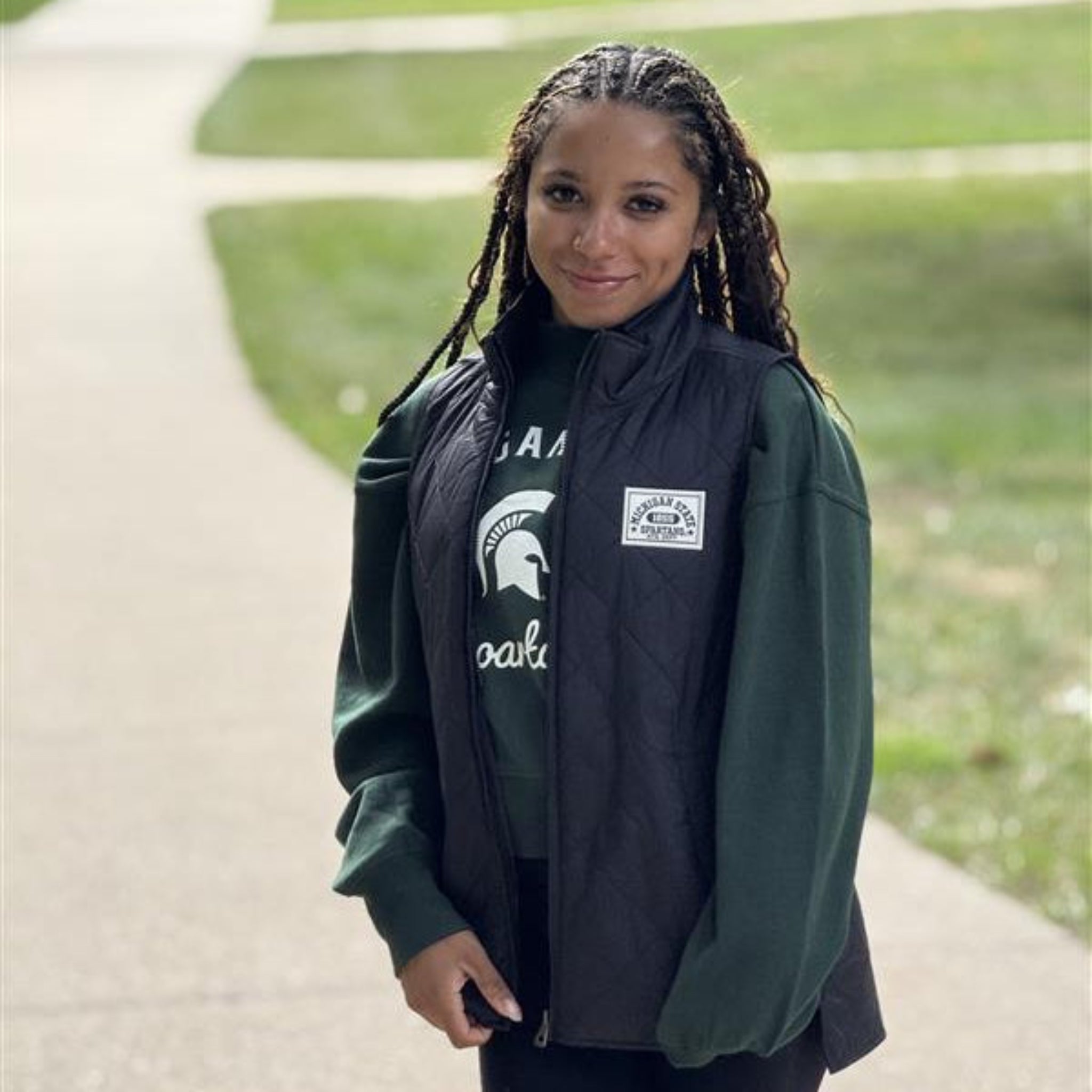 Person wearing a black vest with Michigan State patched over a green jacket with a logo, standing on a path outdoors.
