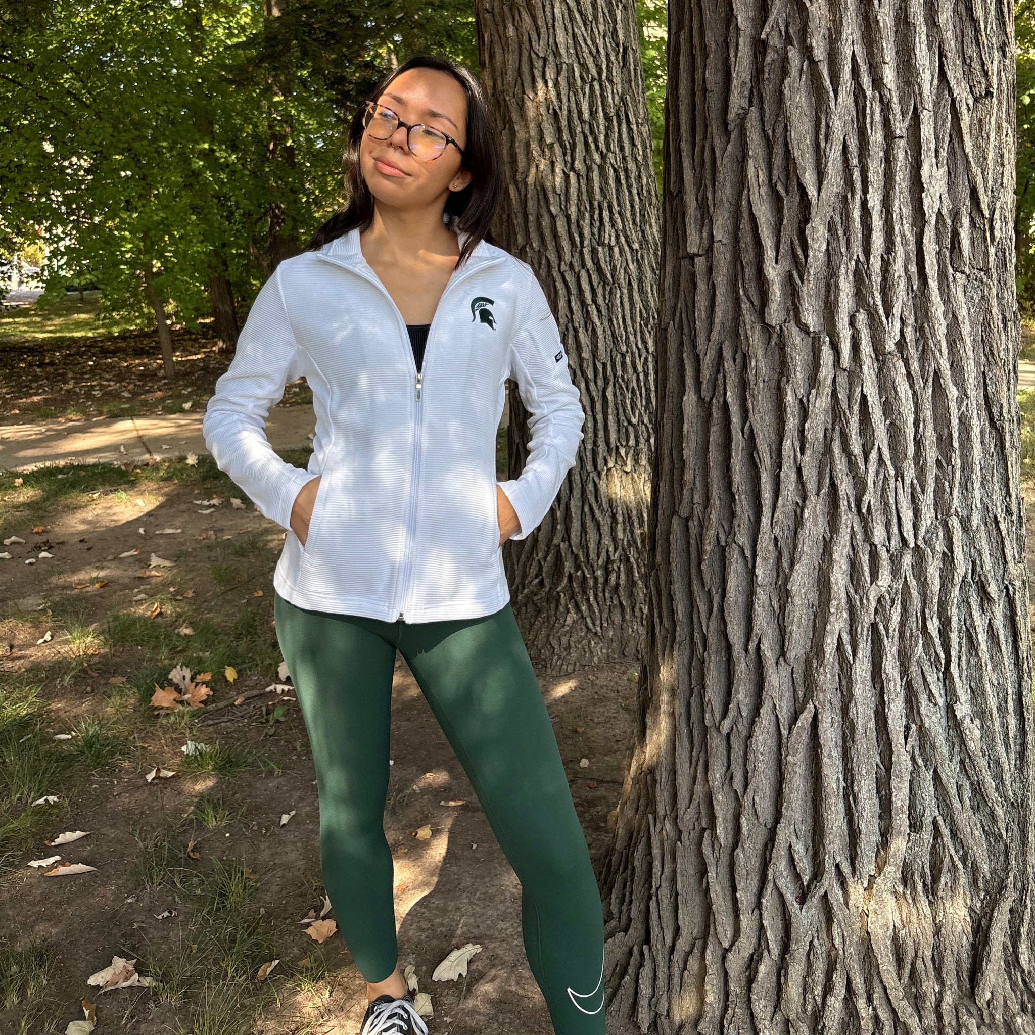 Person in a white striped long-sleeve full-zip jacket with a green embroidered Spartan helmet logo on the left upper chest and pockets at the bottom on either side. 