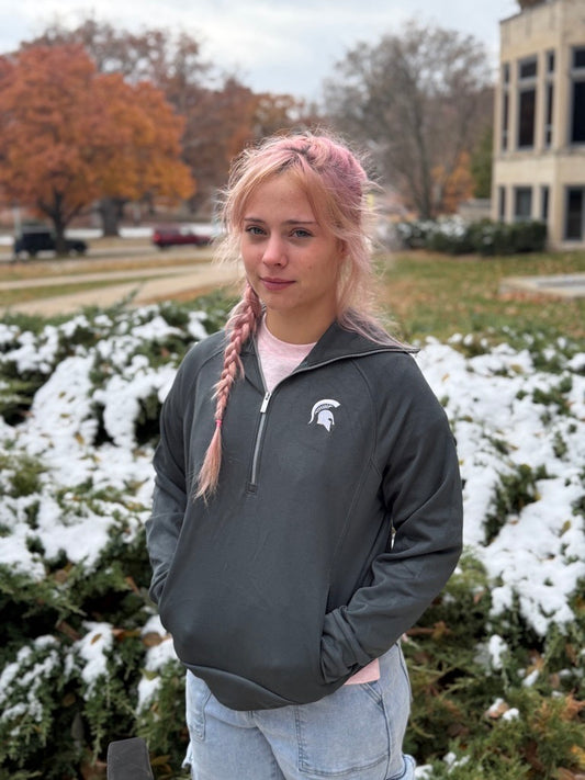Woman, standing outdoors, wearing a gray quarter-zip with a white Spartan Helmet logo on the left upper chest and an extended collar. 