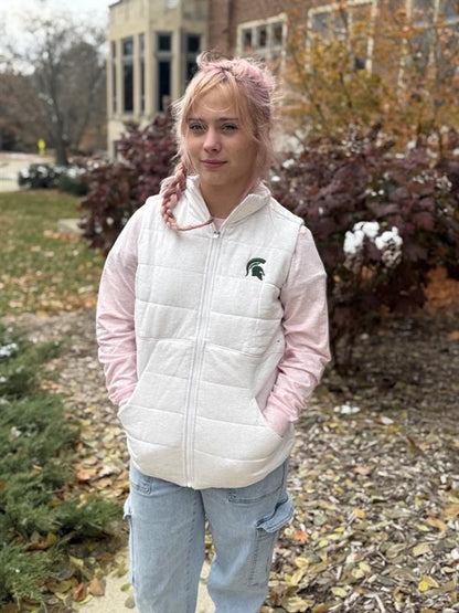 Woman, standing outdoors, wearing an off white quilted vest with horizontal stitching, a front zipper, and a green Spartan helmet logo on the left upper chest. 
