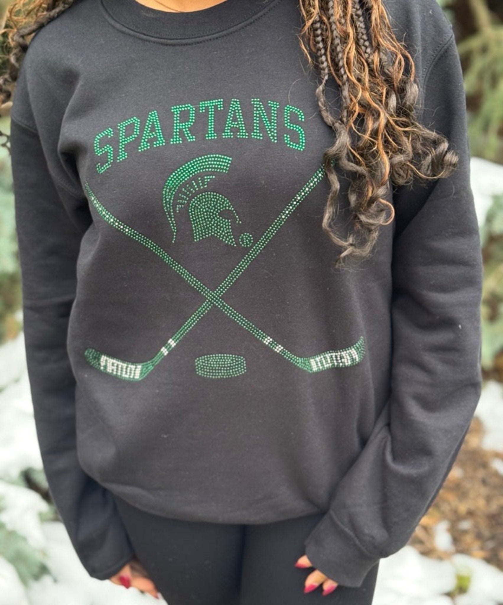 Close up of a hockey themed crewneck. The front of the crewneck displays the word “SPARTANS” in sparkling green stones, along with the iconic Spartan helmet logo, two crossed hockey sticks, and a puck
