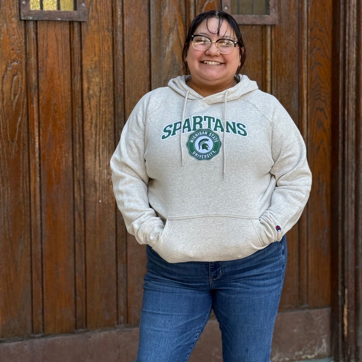 Woman wearing a cream hoodie with "Spartans" and the Michigan State University seal logo with the Spartan Helmet logo inside on the chest, both in green and white. 