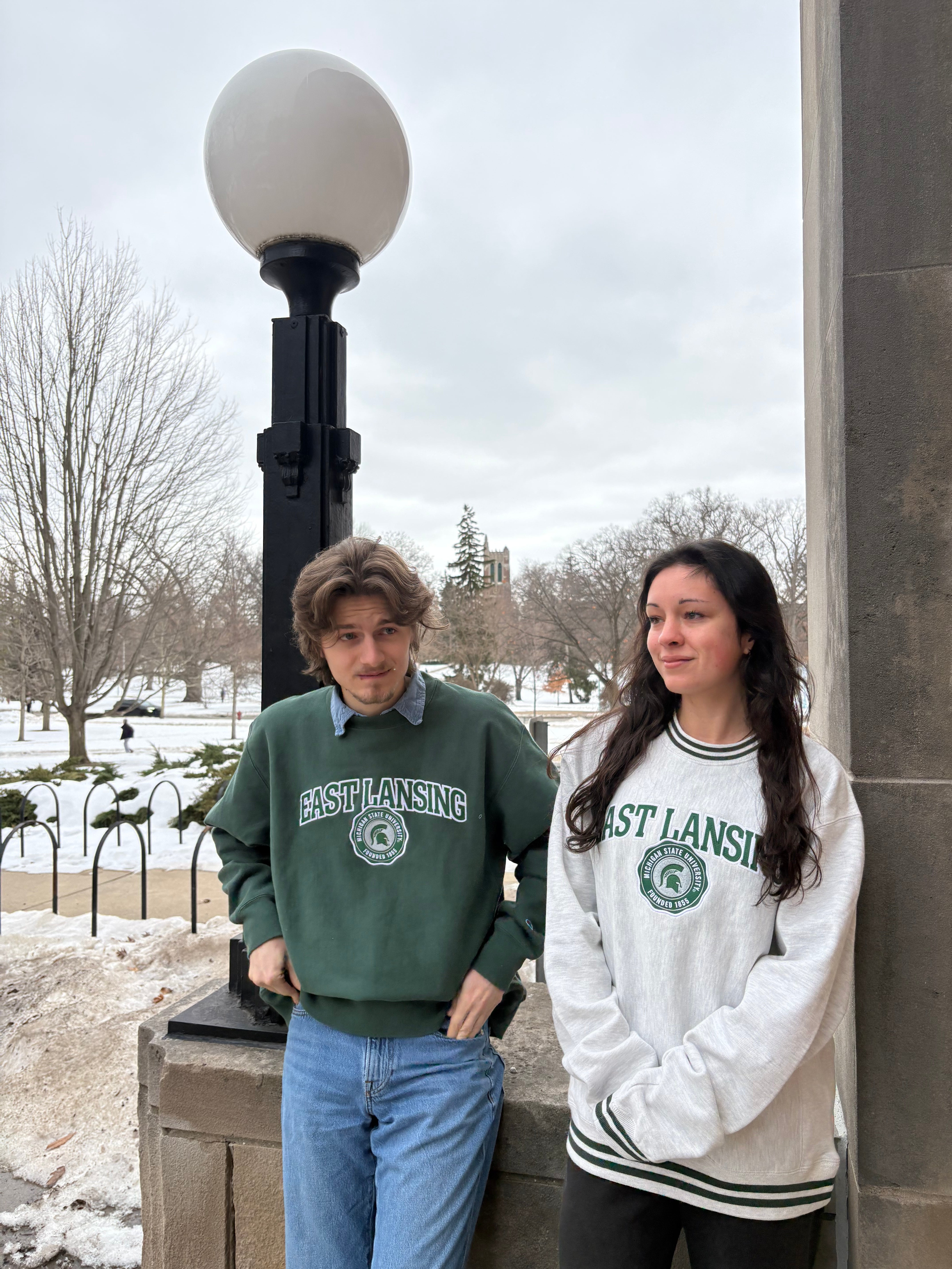 Man and woman standing next to each other outdoors. The man on the left side of the screen is wearing a green crewneck with "East Lansing" written on the front and the Michigan State University seal logo underneath, both in green and white.  The woman on the ride side of the screen is wearing a white crewneck with "East Lansing" written on the front and the Michigan State University seal logo underneath, both in green and white. 