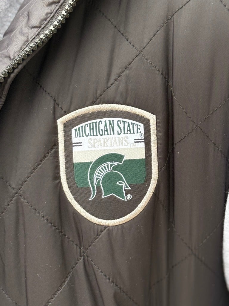 Close up of the a black quilted vest, showing the  an official Spartans logo patch on the upper left chest. 