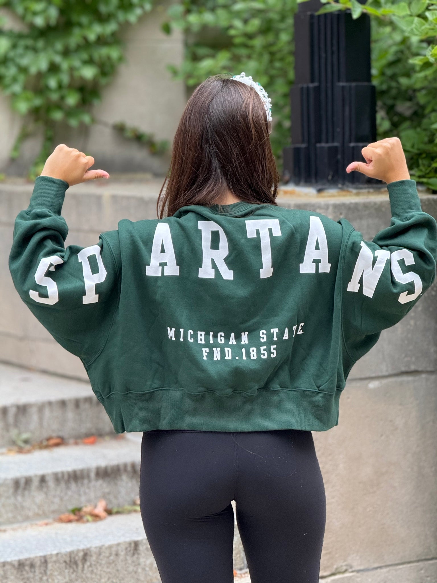 Woman standing outdoors, showing off the back of a green cropped crewneck sweatshirt with a "Spartans" wordmark across the arms and upper back and a "Michigan State FND. 1855" text below it, both printed on in white. 