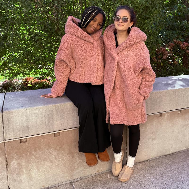 Two woman modeling the Jill Martin pink loungers. The woman on the left of the image is sitting, wearing the cropped pink lounger with a hood. The woman on the left is standing, wearing the long pink lounger with her hand in the pocket. 