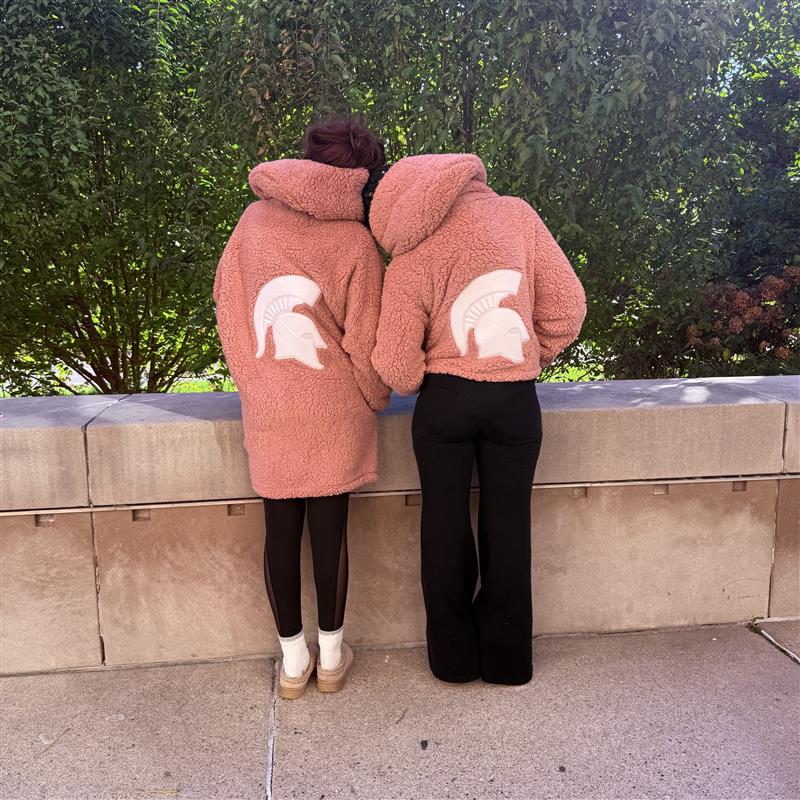 Two woman standing outdoors, modeling the back of the Jill Martin pink loungers. The woman on the left is wearing the long pink lounger with a white Spartan Helmet logo centered on the back. The women on the right is wearing the cropped pink lounger with a white Spartan Helmet logo centered on the back. 