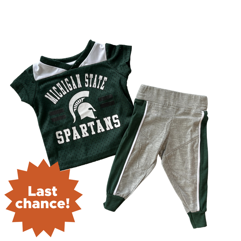 A toddler or infant sized green football jersey with Michigan State Spartans and the MSU helmet logo written on the front. Grey sweatpants with green lines going down the side of the pant legs accompany the jersey. In the lower left is an orange sunburst with the words "Last chance!'