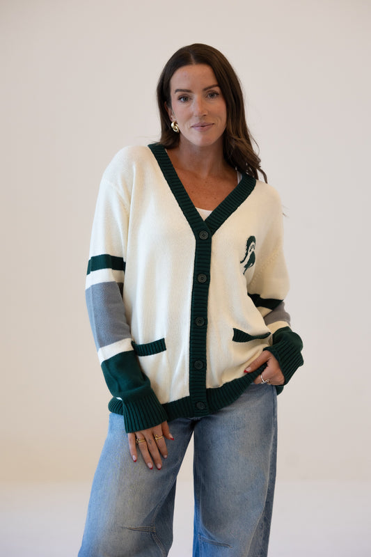 Woman wearing a cream Michigan State University cardigan with dark green trim, striped sleeves, and an embroidered Spartan logo, paired with blue jeans.