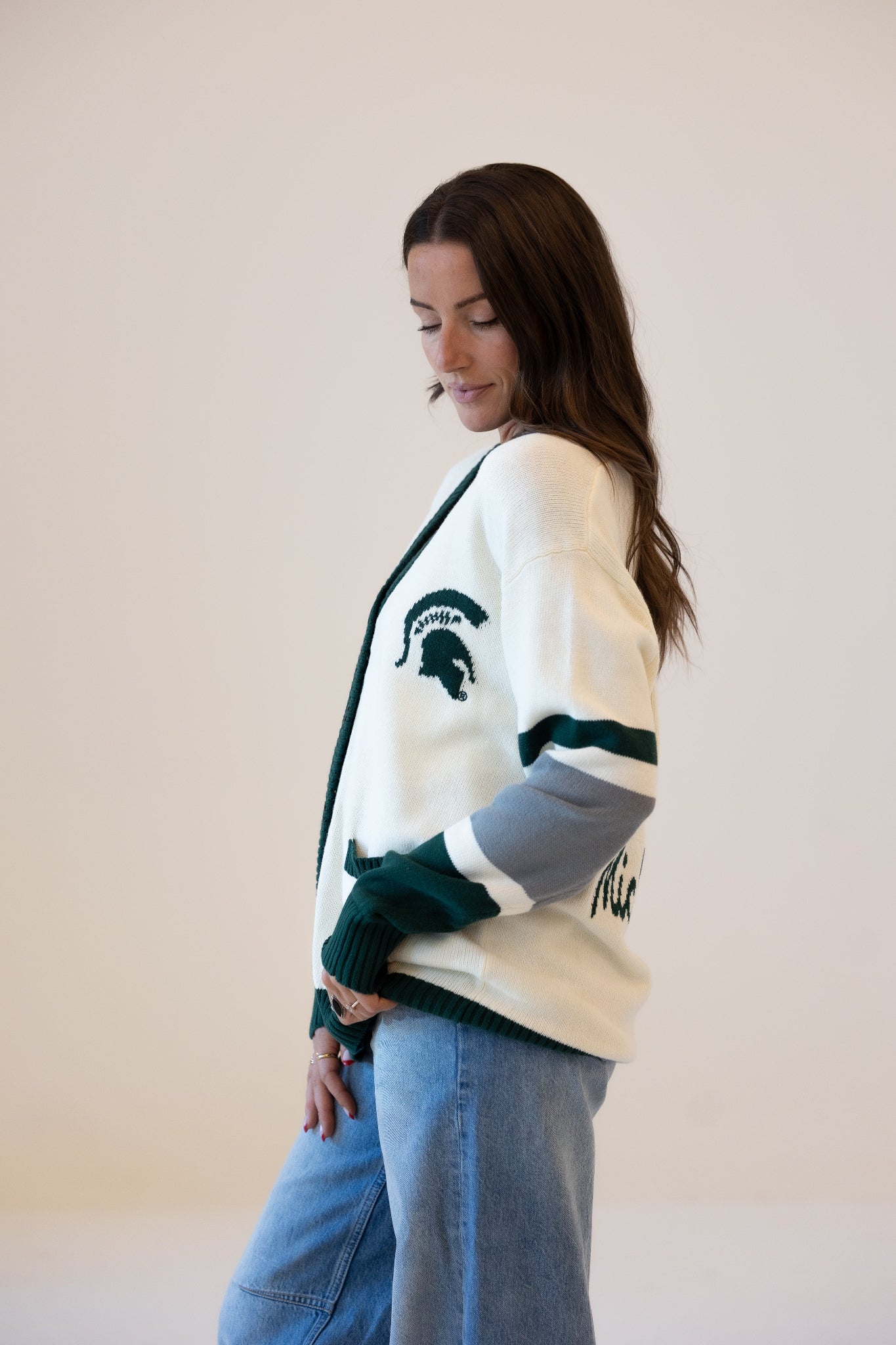 Woman wearing a cream Michigan State University cardigan with dark green trim, a Spartan helmet logo, and green, white, and gray striped sleeves, paired with blue jeans.