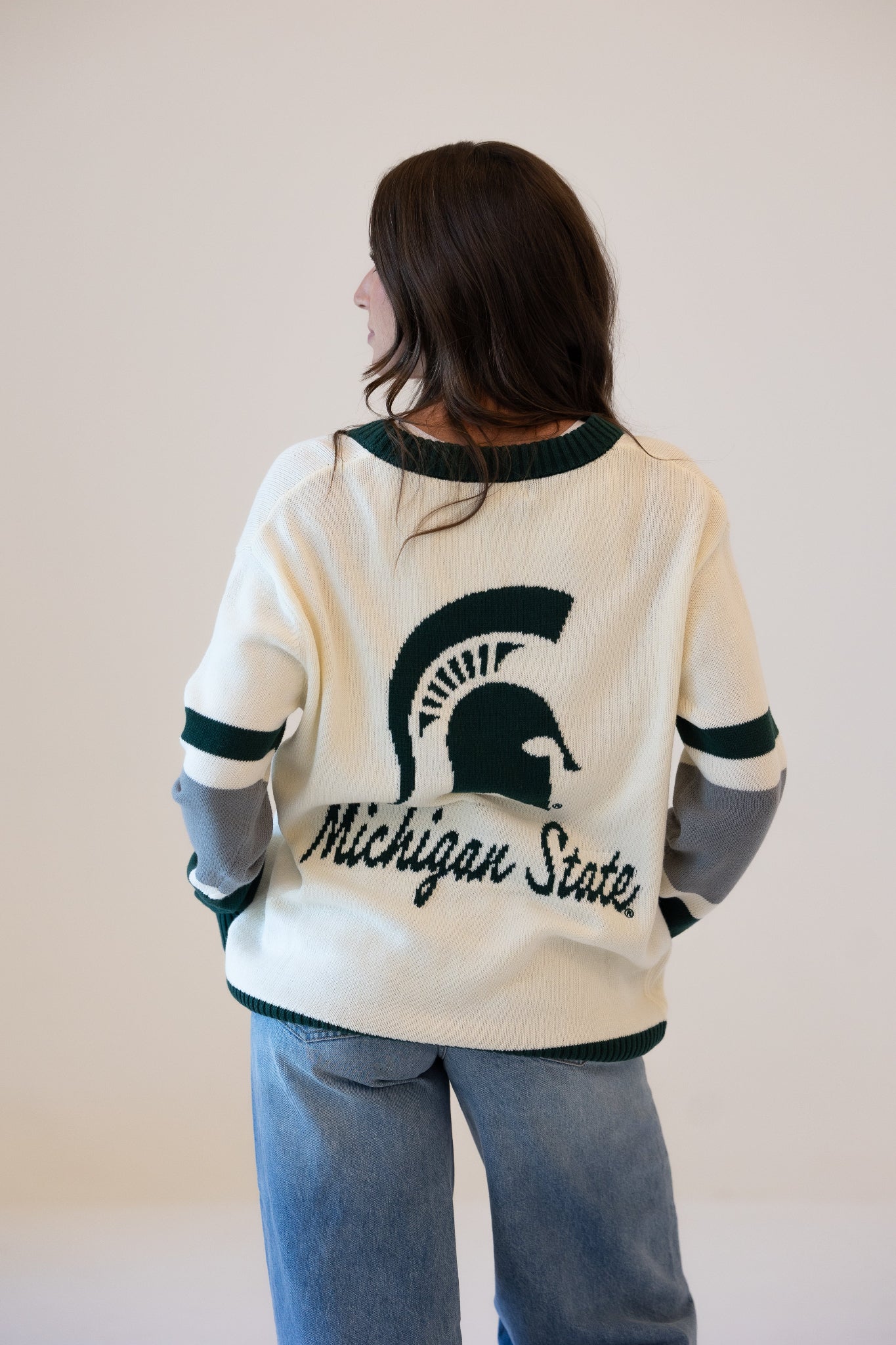 Woman shown from the back wearing a cream Michigan State University cardigan with dark green trim, a large Spartan helmet graphic, “Michigan State” lettering, and green, white, and gray striped sleeves, paired with blue jeans.