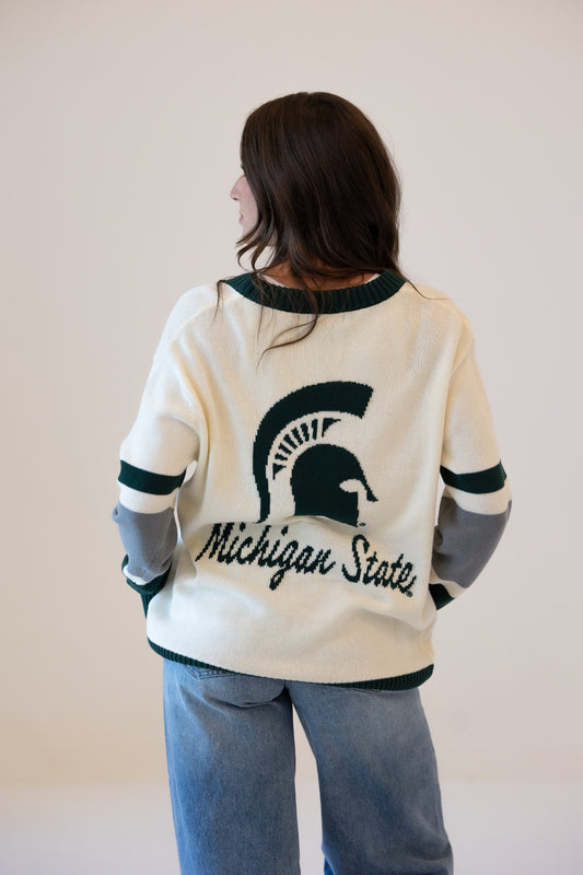 Woman shown from the back wearing a cream Michigan State University cardigan with dark green trim, a large Spartan helmet graphic, “Michigan State” lettering, and green, white, and gray striped sleeves, paired with blue jeans.