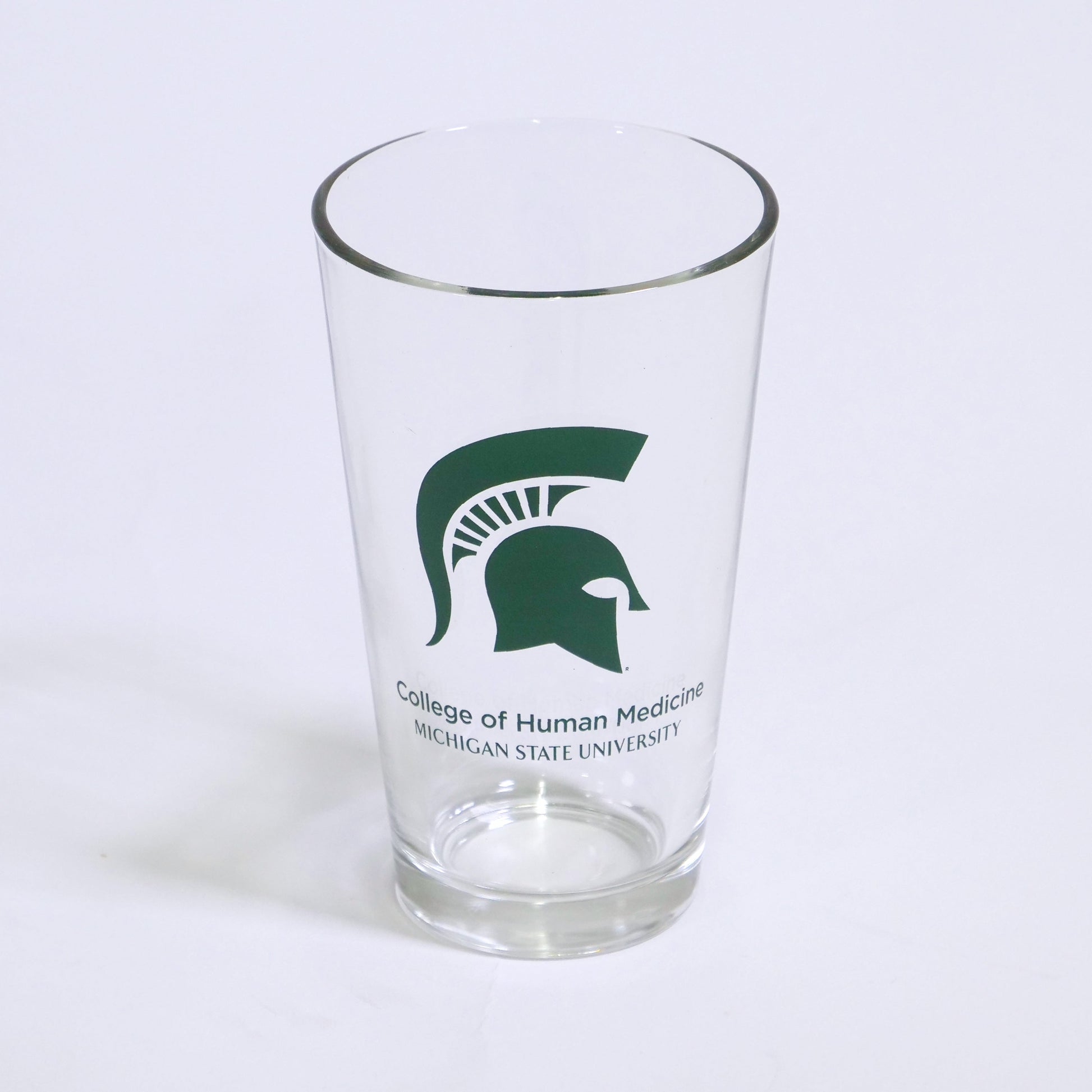 Clear tapered pint glass with a large green Spartan helmet logo printed on the center. Below the Spartan Helmet logo, green text that reads "College of Human Medicine Michigan State University" on two lines.