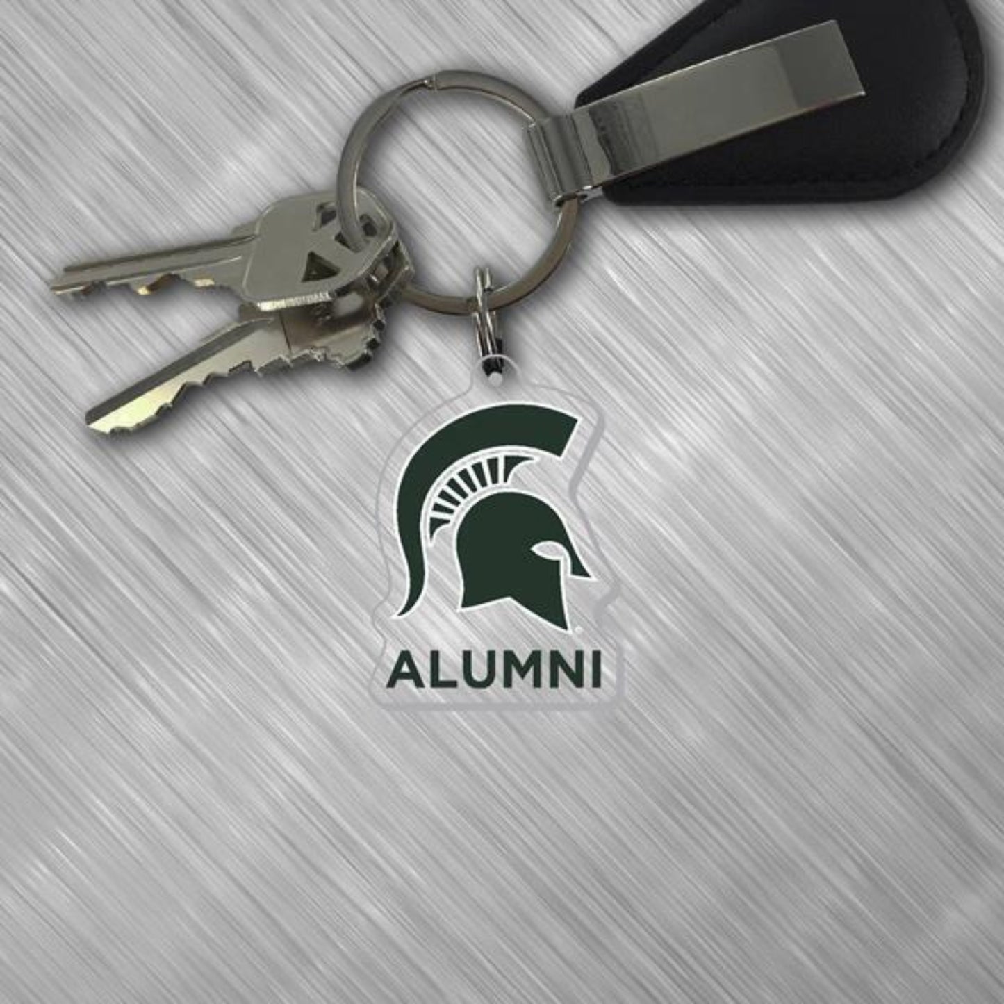 Michigan State University Alumni acrylic keychain attached to a metal key ring with keys. The clear keychain features the green Spartan helmet logo above the word “ALUMNI” in bold lettering. Displayed on a light gray textured surface with a black key fob attached to the ring.