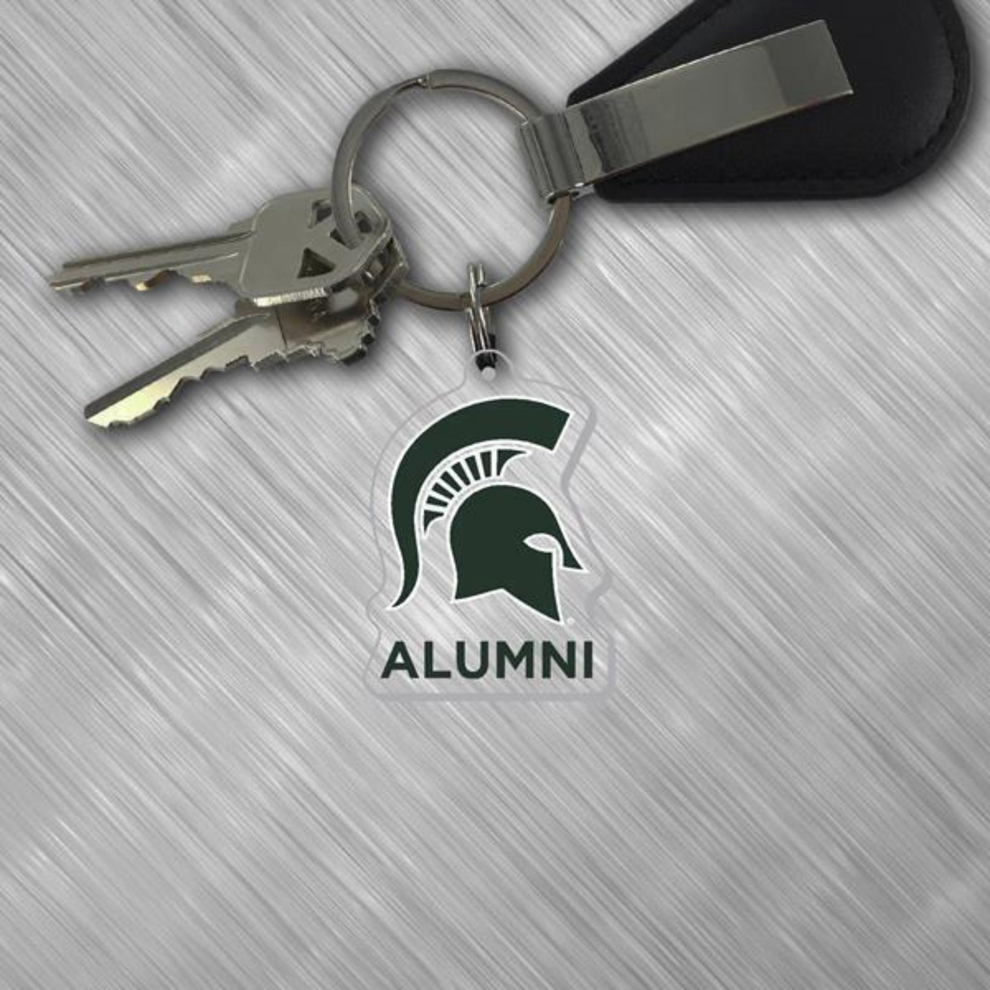 Michigan State University Alumni acrylic keychain attached to a metal key ring with keys. The clear keychain features the green Spartan helmet logo above the word “ALUMNI” in bold lettering. Displayed on a light gray textured surface with a black key fob attached to the ring.