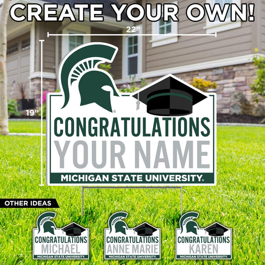 A white customizable yard sign with 'CONGRATULATIONS' text in green, a black graduation cap, and a Michigan State Spartan helmet in green at the top. At the bottom is a green stripe with the words 'Michigan State University' in white.