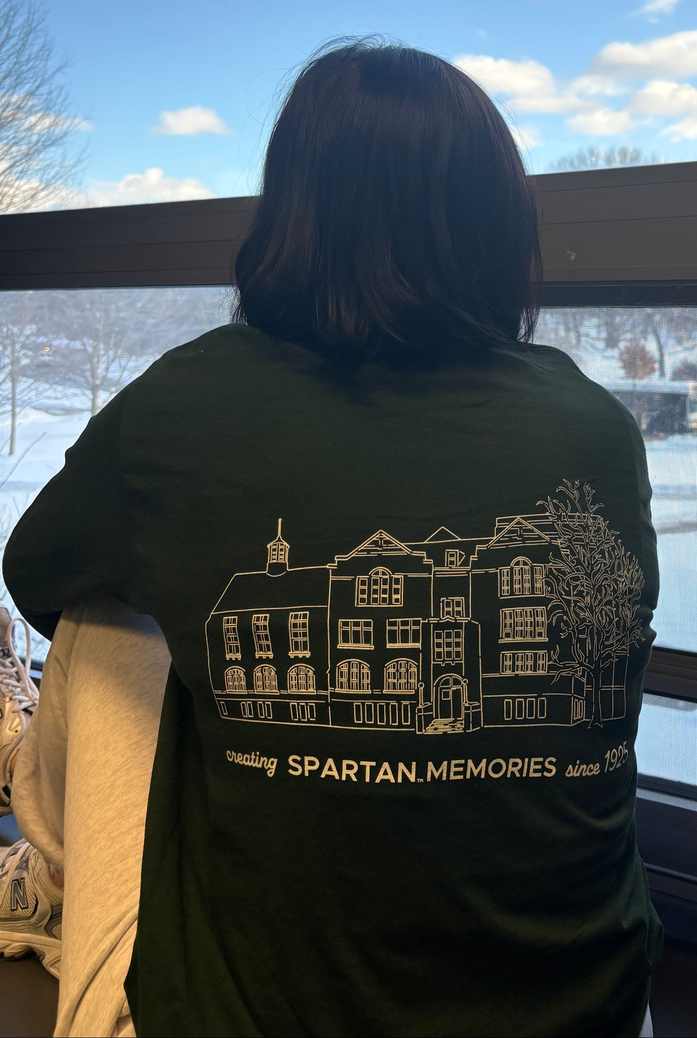 Person wearing a dark green long sleeve shirt with text of "Creating Spartan memories since 1925" and graphic of MSU Union building with trees, standing by a window with a snowy landscape outside.