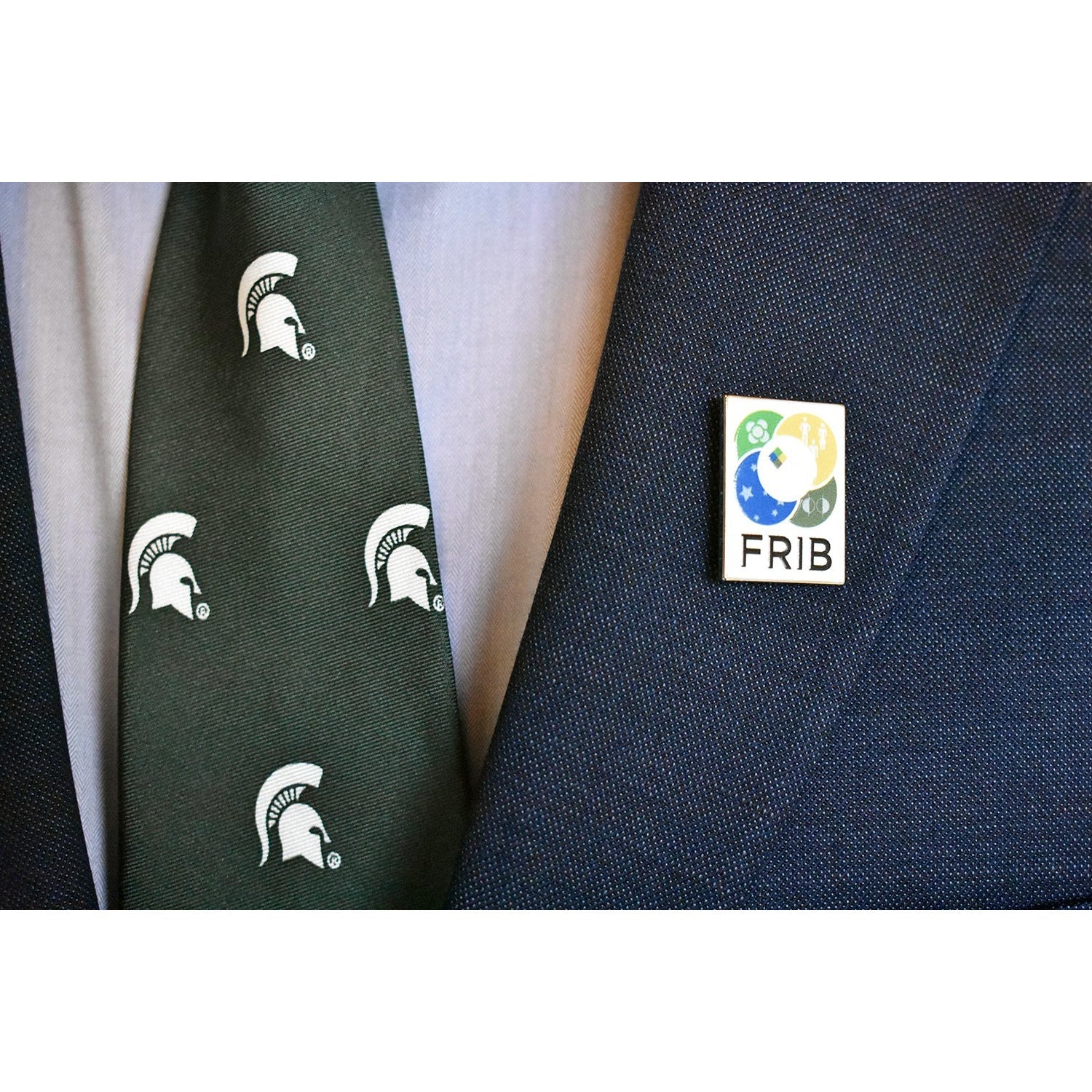 Close-up of a navy suit with the FRIB pin attached to the lapel. The wearer has a green tie featuring white Spartan helmets on as well.