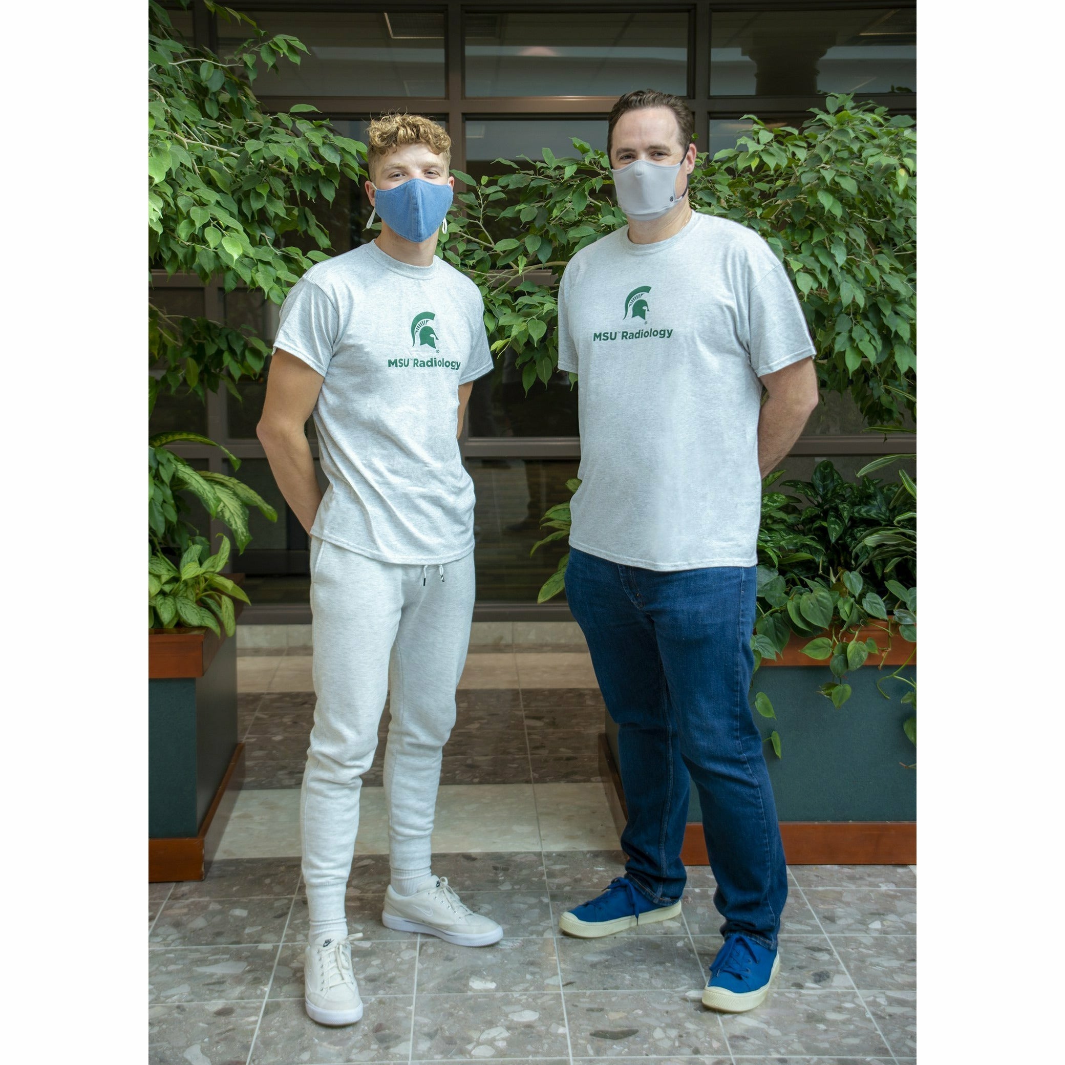 Two men stand side-by-side within the gardens at a slight angle to show the gray t-shirts from the side.