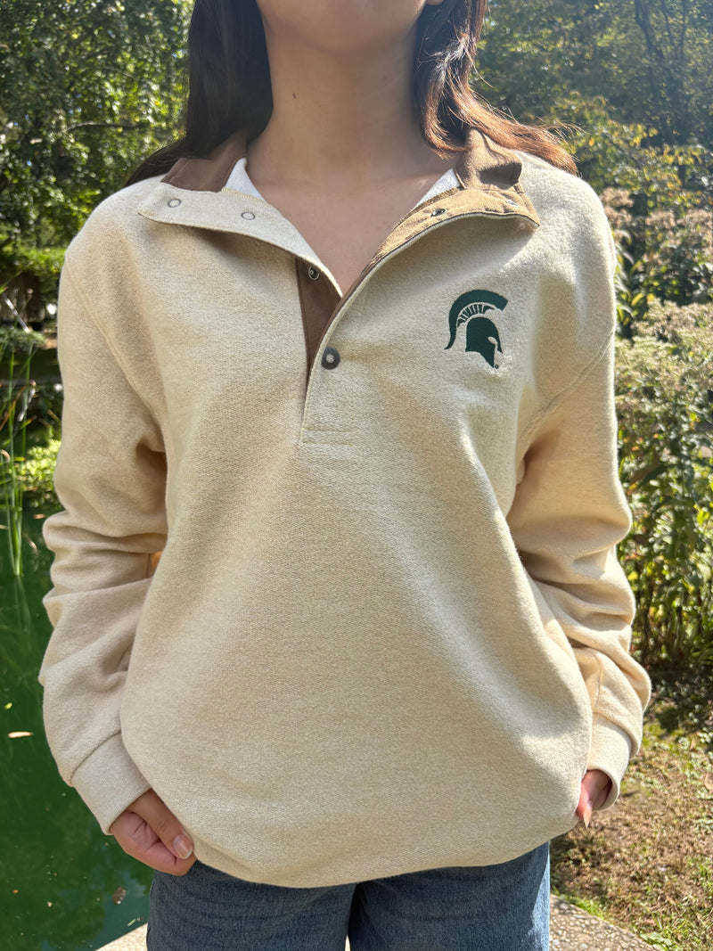 Person wearing a beige jacket with a  green Spartan helmet logo, standing outdoors.