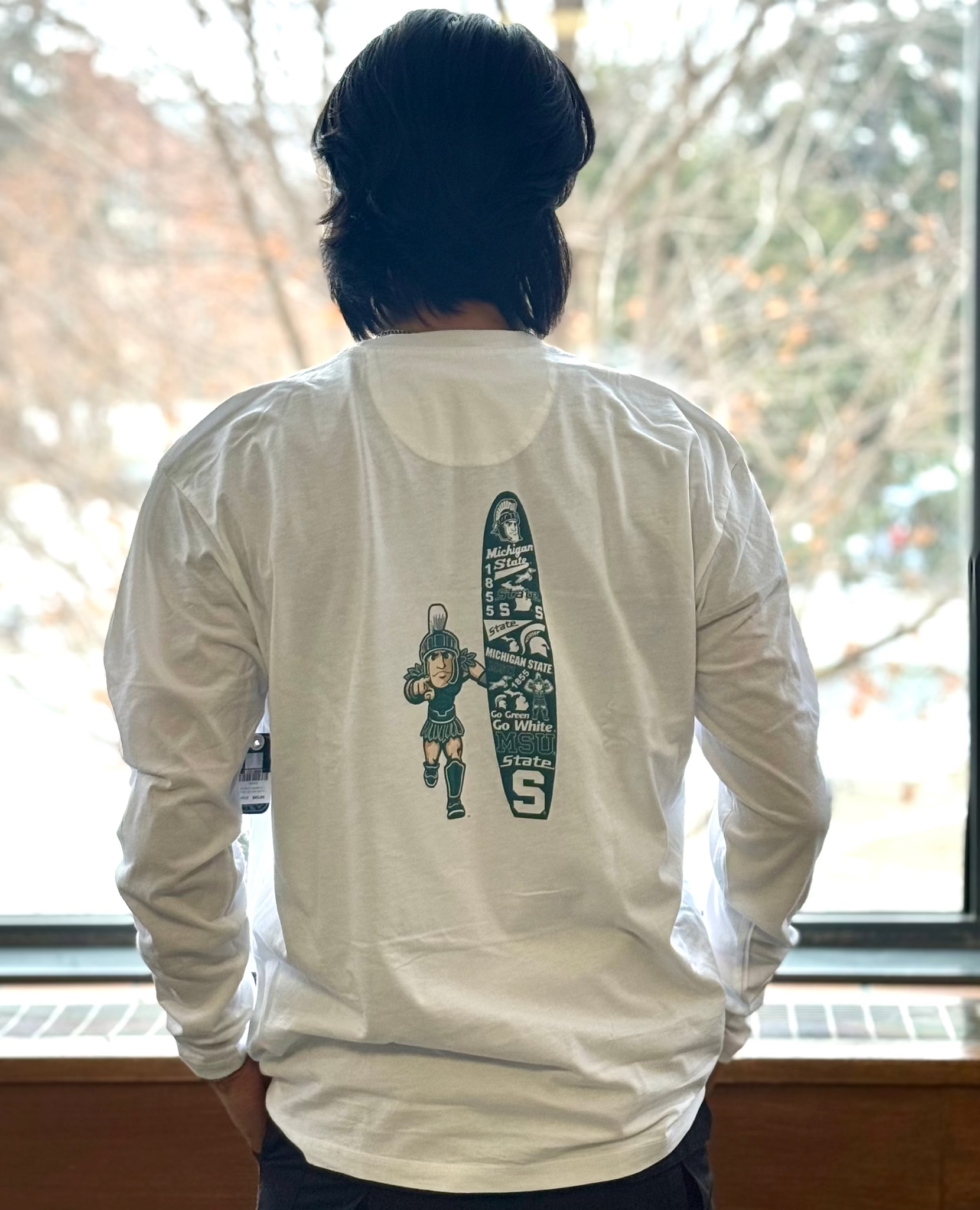Person wearing a white long-sleeve shirt with a screen printed Sparty and green surf board with various Michigan State University logo's on the back, standing in front of a window.