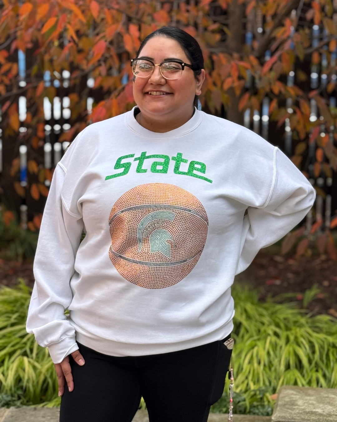 Woman standing outdoors wearing a white long sleeve crewneck sweatshirt with the word "State" written in glittery green text near the top and a rhinestone orange basketball with a Spartan Helmet logo in the middle. 