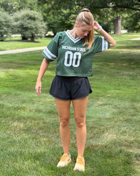 A person stands on grass wearing a green Michigan State jersey with the number 00 and black shorts. The individual is looking down and adjusting their hair. Trees and a walkway are in the background.