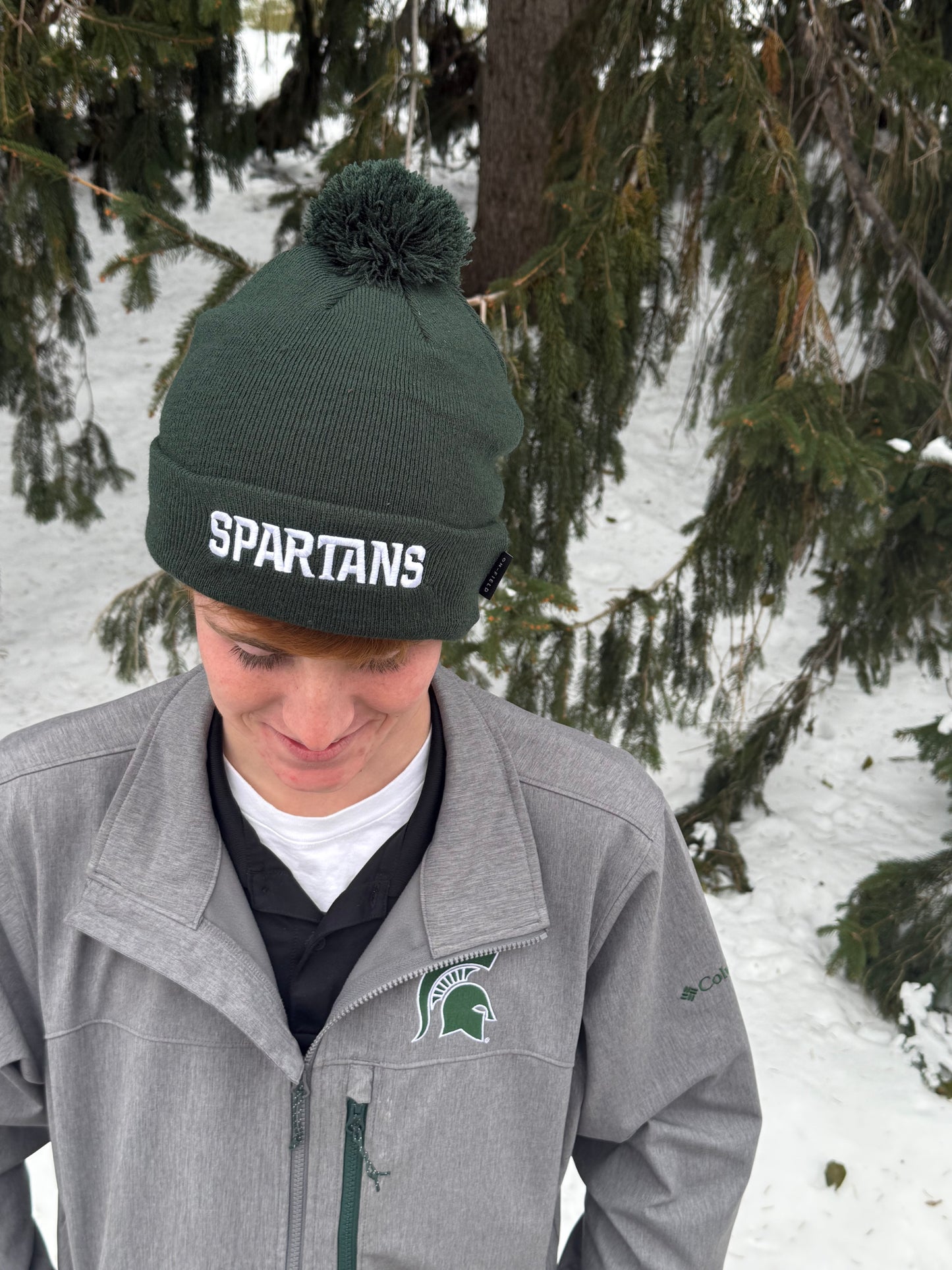 Close up, showing the top of a green cuffed beanie with a green pom at the top and a white Spartans wordmark embroidered on the cuff. 