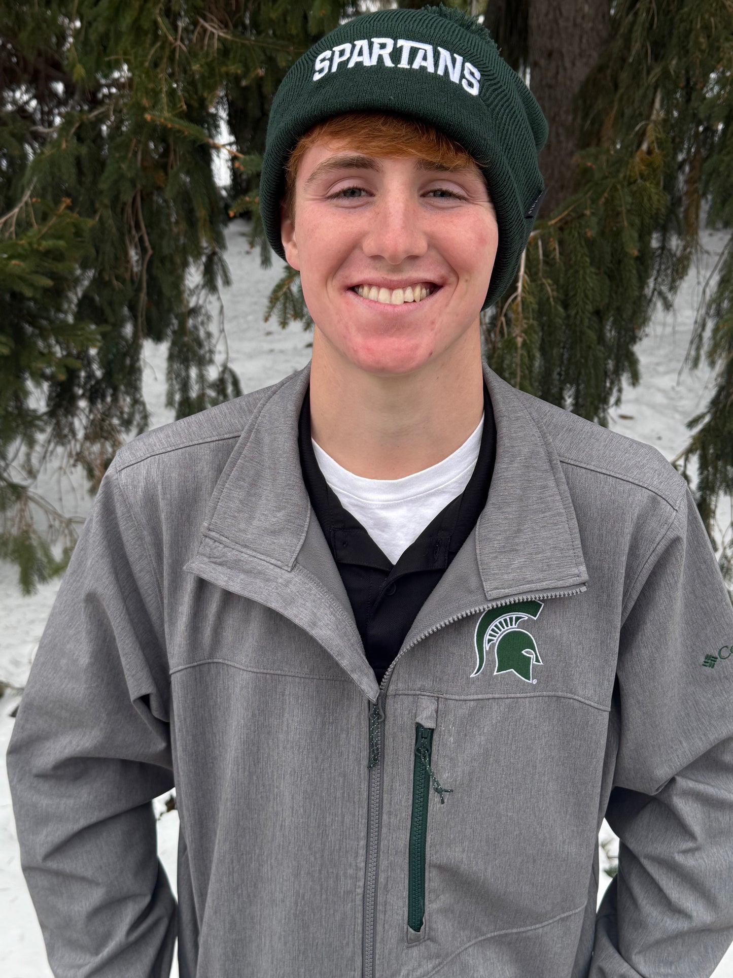 Man standing outdoors, wearing a green cuffed beanie with a green pom at the top and a white Spartans wordmark embroidered on the cuff. 