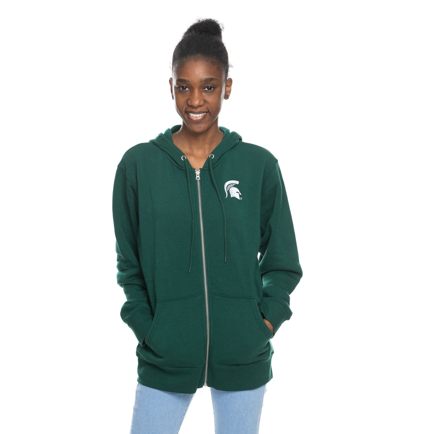 Woman with hands in pockets wearing a green full-zip hoodie with a white Spartan Helmet logo at left chest.